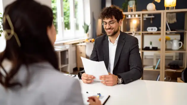 Two people are in an interview setting. One person, who is seated at a table, is reviewing a document.